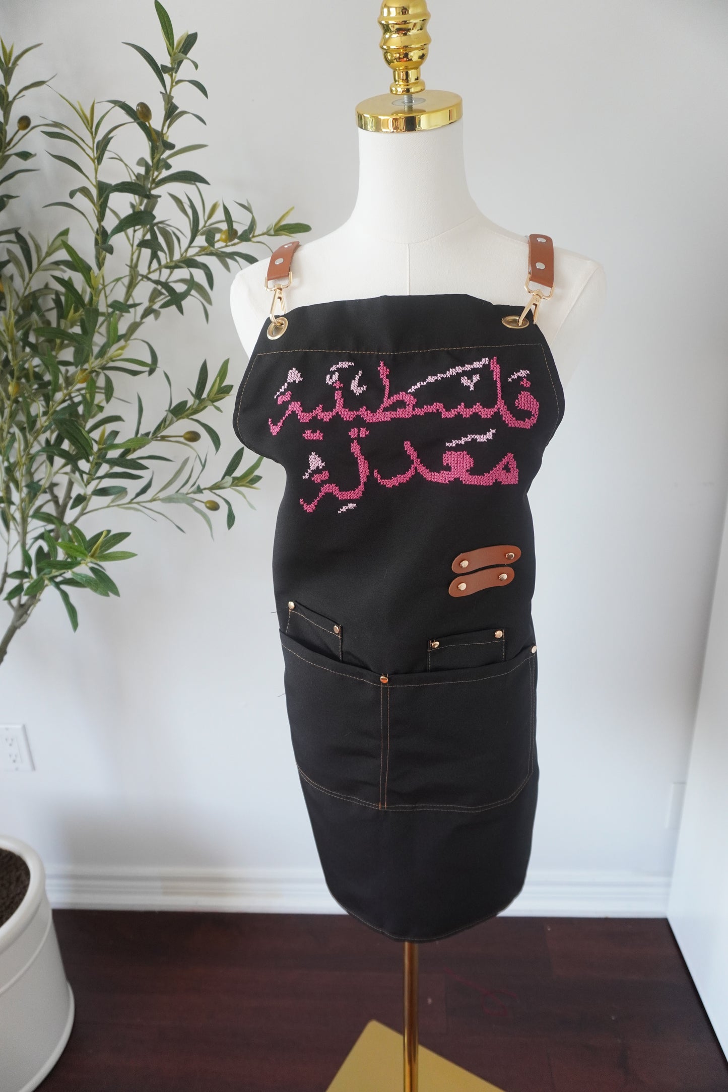 Adjustable apron with grey straps, featuring hand-embroidered Palestinian Felestenea Madala design in pink gradients, crafted with traditional tatreez techniques. Includes deep front pockets and a flattering waistline.