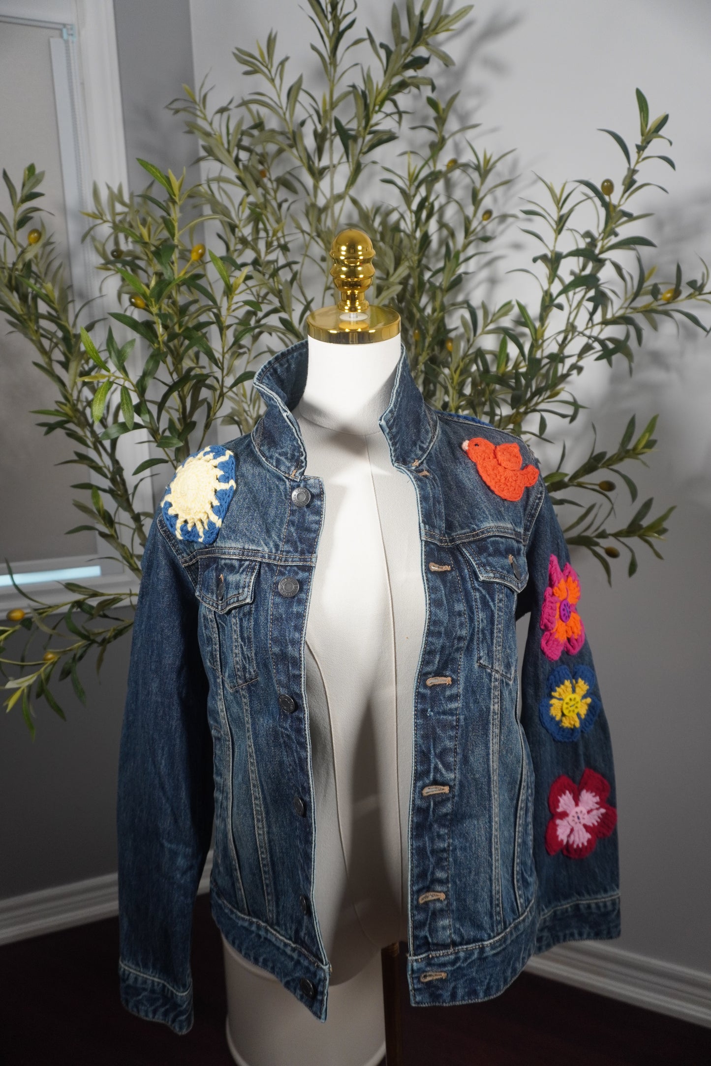 A small-sized denim jacket featuring colorful crochet squares with buttons on the back, flower and button embellishments on the sleeves, and a decorative bird on the front, showcasing one-of-a-kind artistry by a talented artist from Lebanon.
