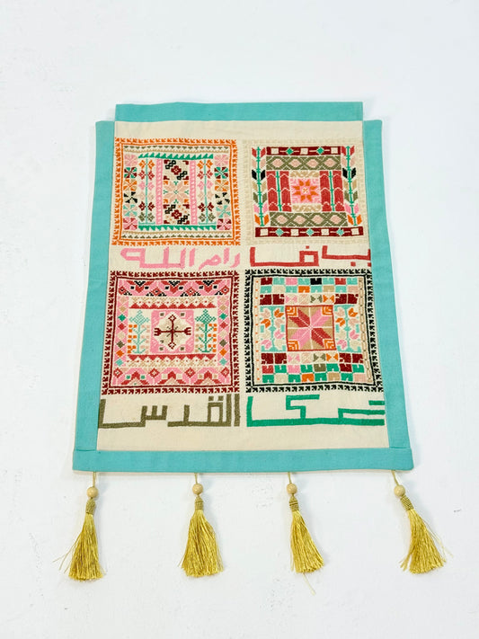 Handmade tatreez wall hanging with embroidered Palestinian city names and colorful geometric motifs on fabric, crafted by women artisans.