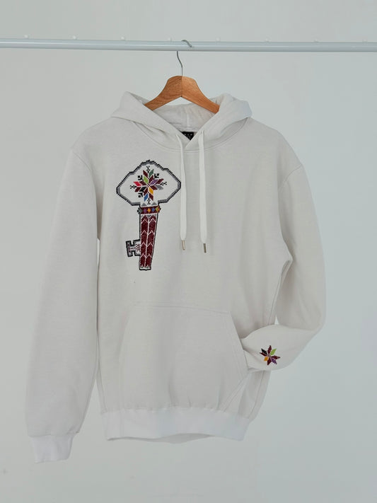 White cotton hoodie with hand-embroidered Palestinian key of return motif (مفتاح العودة) on the left chest, displayed on hanger, handmade by women artisans