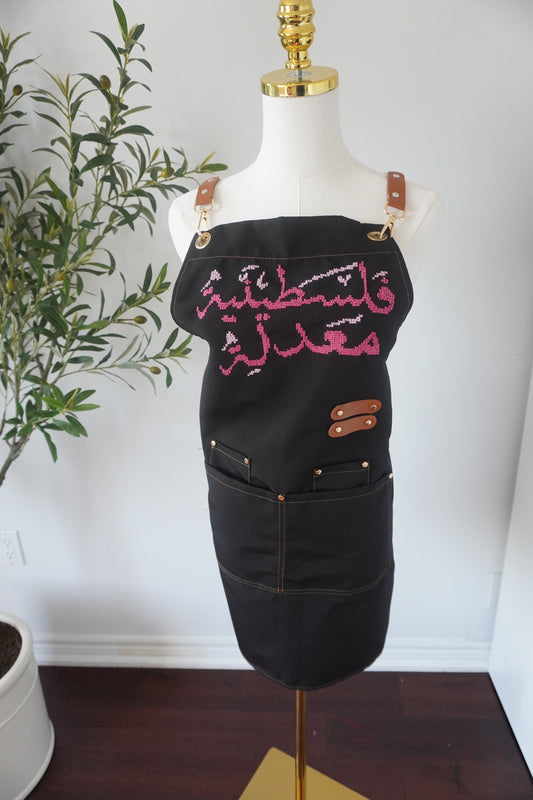 Adjustable apron with grey straps, featuring hand-embroidered Palestinian Felestenea Madala design in pink gradients, crafted with traditional tatreez techniques. Includes deep front pockets and a flattering waistline.