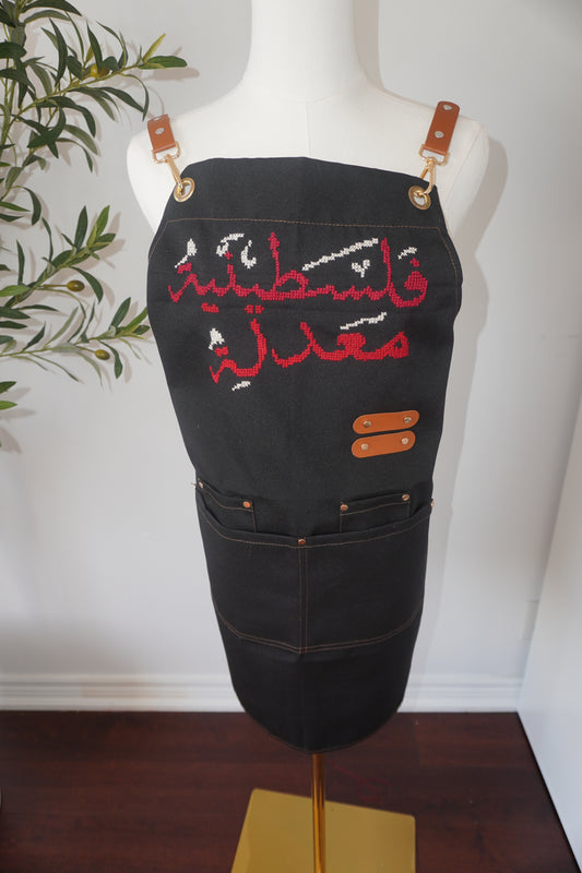 Adjustable apron with grey straps, featuring hand-embroidered Palestinian Felestenea Madala design in red and white, crafted with traditional tatreez techniques. Includes deep front pockets and a flattering waistline.
