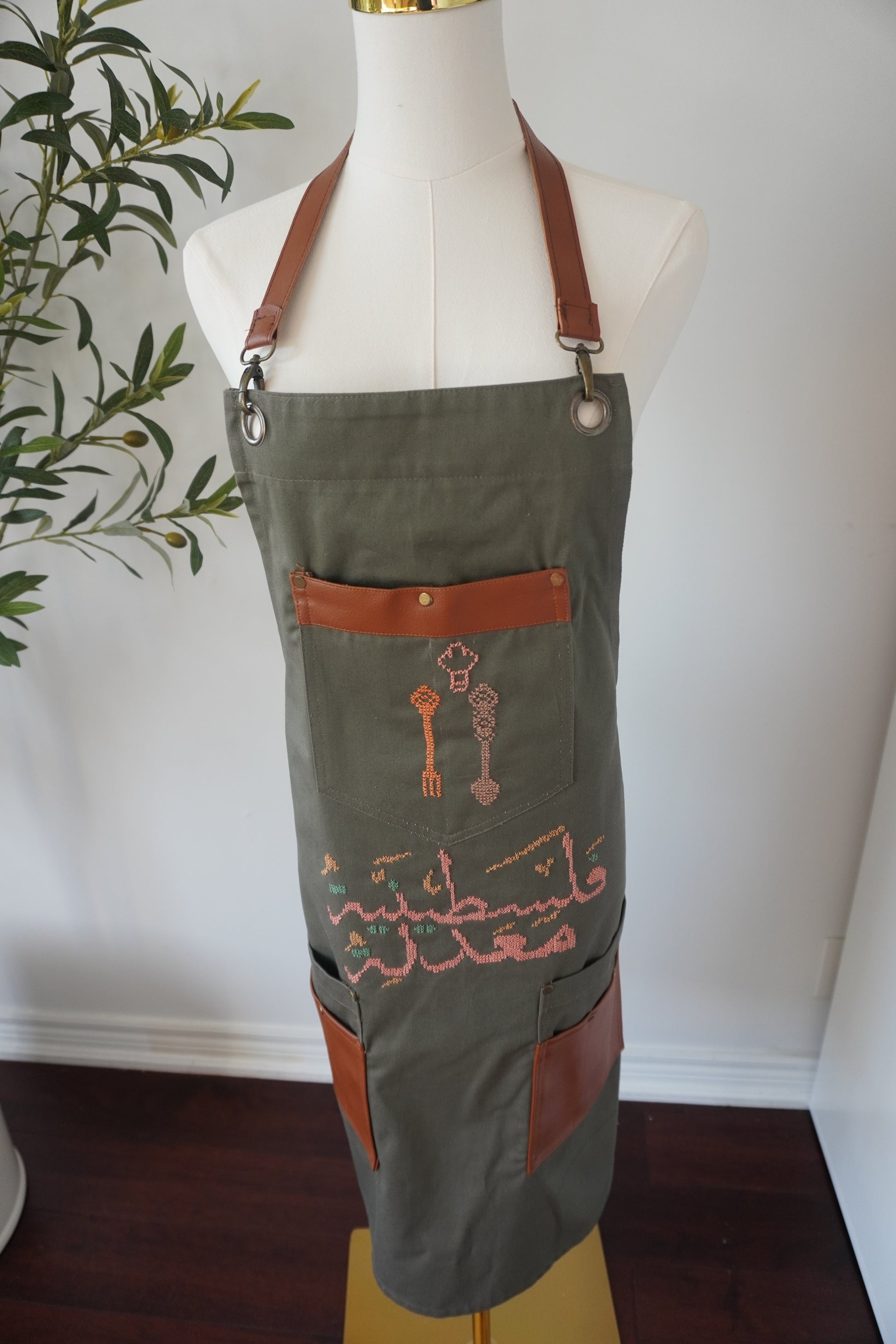 Olive green apron featuring hand-embroidered Palestinian motif with peach and brown spoons, designed to be super beautiful and cute.