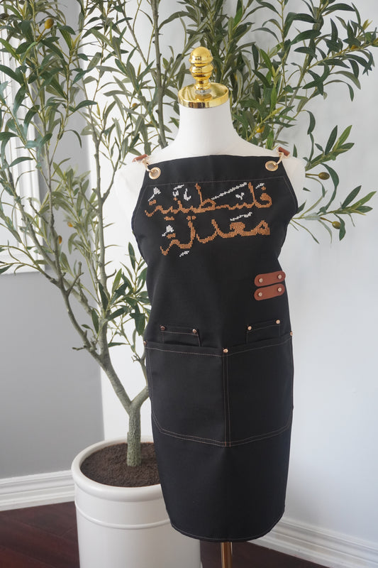 Adjustable apron with grey straps, featuring hand-embroidered Palestinian modified design in cumon color, crafted with traditional tatreez techniques. Includes deep front pockets and a flattering waistline.