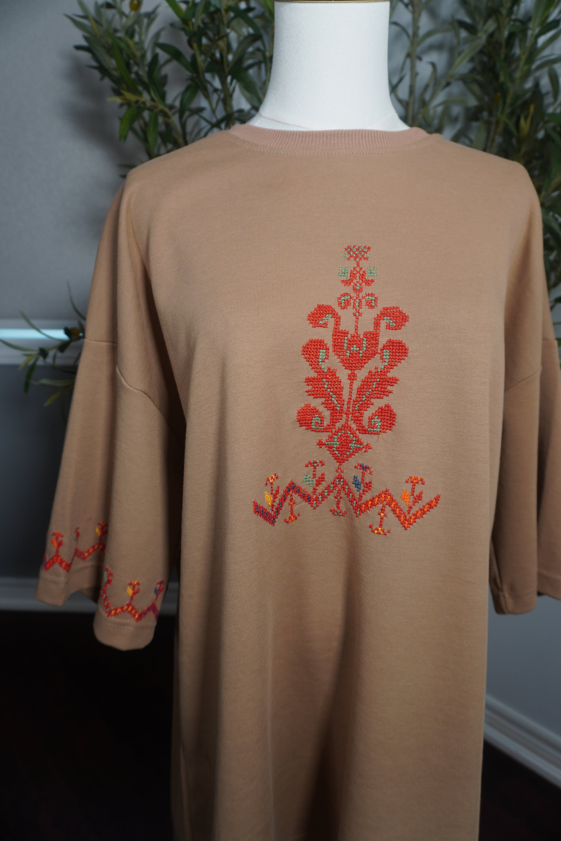 A unique oversized beige t-shirt featuring intricate Palestinian tatreez embroidery in vibrant red, with accents of green, orange, and light green, and a decorative bracelet on one sleeve, showcasing the artistry of hand-stitching