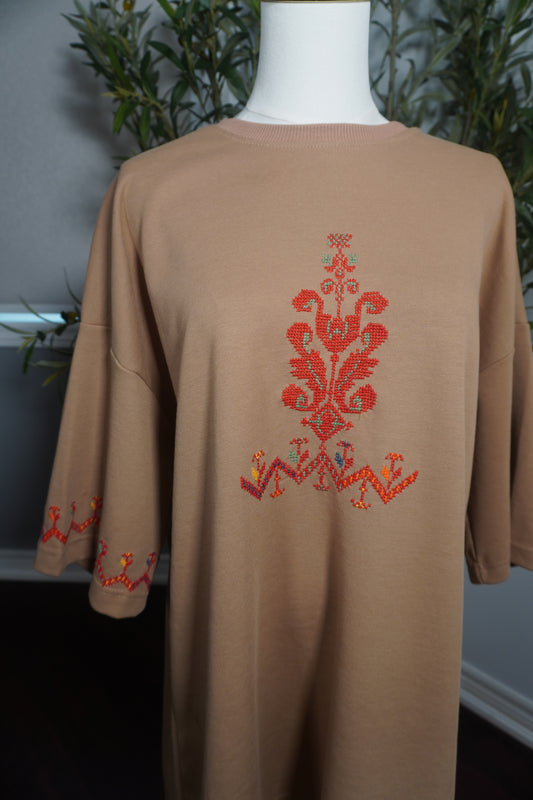 A unique oversized beige t-shirt featuring intricate Palestinian tatreez embroidery in vibrant red, with accents of green, orange, and light green, and a decorative bracelet on one sleeve, showcasing the artistry of hand-stitching