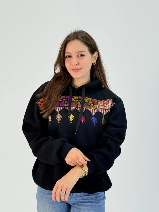 Black cotton hoodie with colorful hand-embroidered Palestinian shepherd and floral tatreez pattern across the chest, modeled with jeans, handmade by women artisans