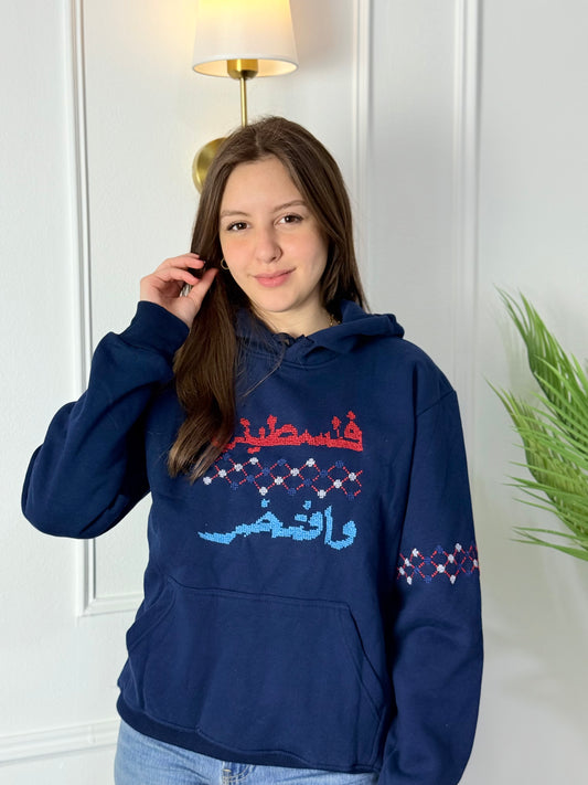 Navy blue cotton hoodie with red and blue hand-stitched Palestinian embroidery saying “فلسطيني وافتخر” (Palestinian and Proud), modeled by a woman, handmade by women artisans