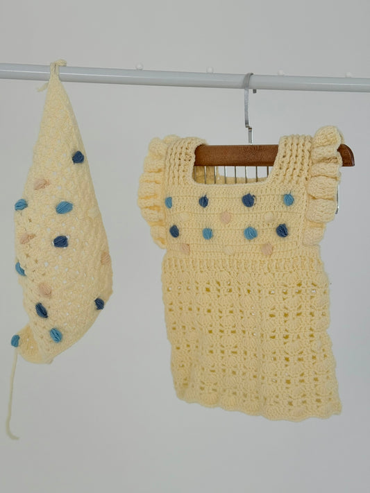 Handmade beige crochet baby dress with matching bandana, featuring blue pom-poms, crafted by women artisans for a soft, breathable fit.

