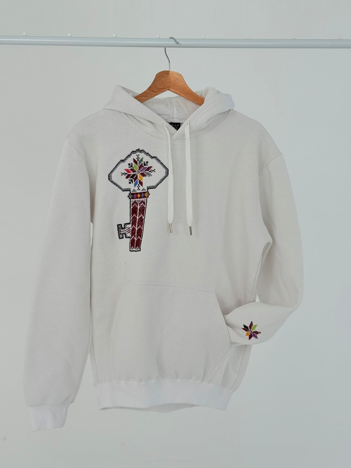 White cotton hoodie with hand-embroidered Palestinian key of return motif (مفتاح العودة) on the left chest, displayed on hanger, handmade by women artisans