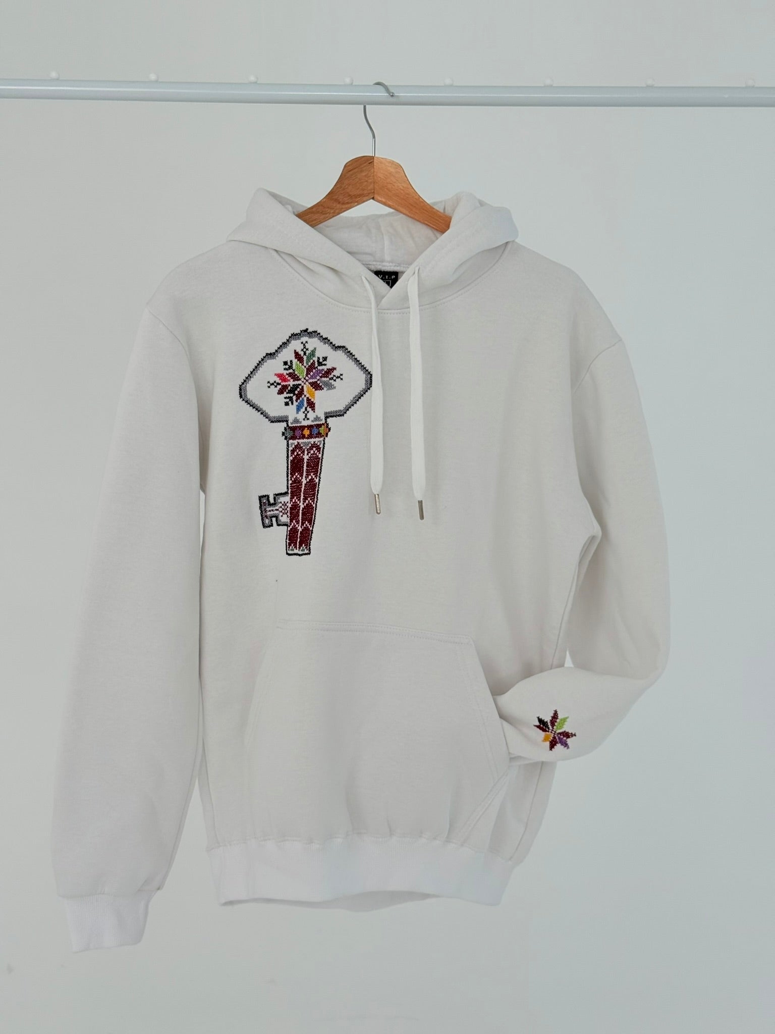 White cotton hoodie with hand-embroidered Palestinian key of return motif (مفتاح العودة) on the left chest, displayed on hanger, handmade by women artisans