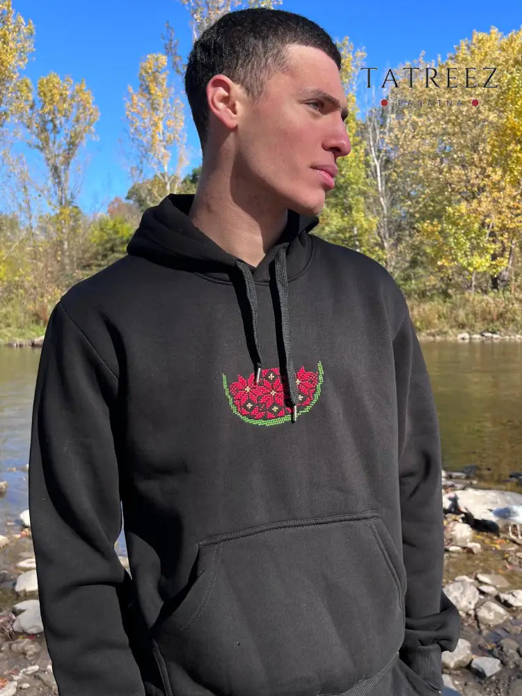 Black cotton hoodie featuring a small hand-stitched watermelon motif at the center of the chest. The design is embroidered in red and green, using traditional Palestinian tatreez to create a half-melon shape composed of simple, flower-like patterns. The hoodie includes a front kangaroo pocket, a drawstring hood, and a relaxed fit.