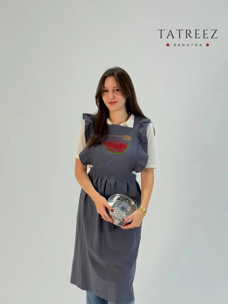 Grey apron with hand-embroidered watermelon and spoon motif, handmade by women artisans in Palestine and Jordan.
