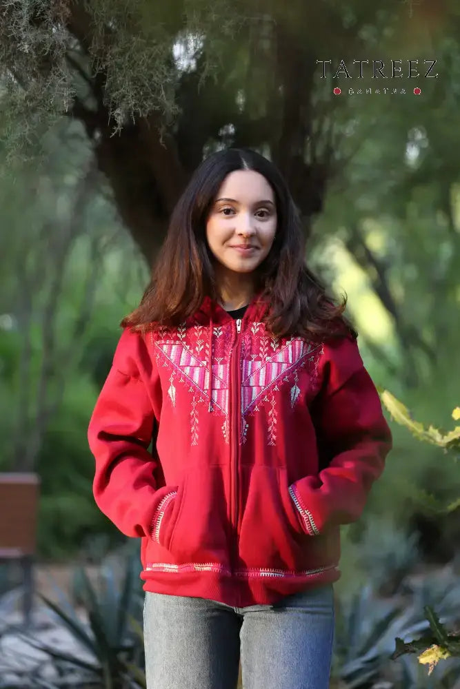 Red 'Laila' sweater with handmade Sanabel embroidery on the front and delicate details on the hoodie edges, crafted by Palestinian refugees in Jordan. Model typically wears a small and is 157 cm tall, perfect for cozy, stylish wear.